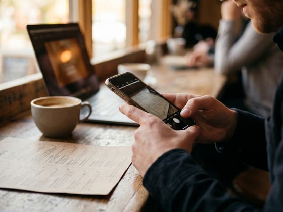 Hands taking a phone picture of a paper menu next to a laptop and a cup of coffee